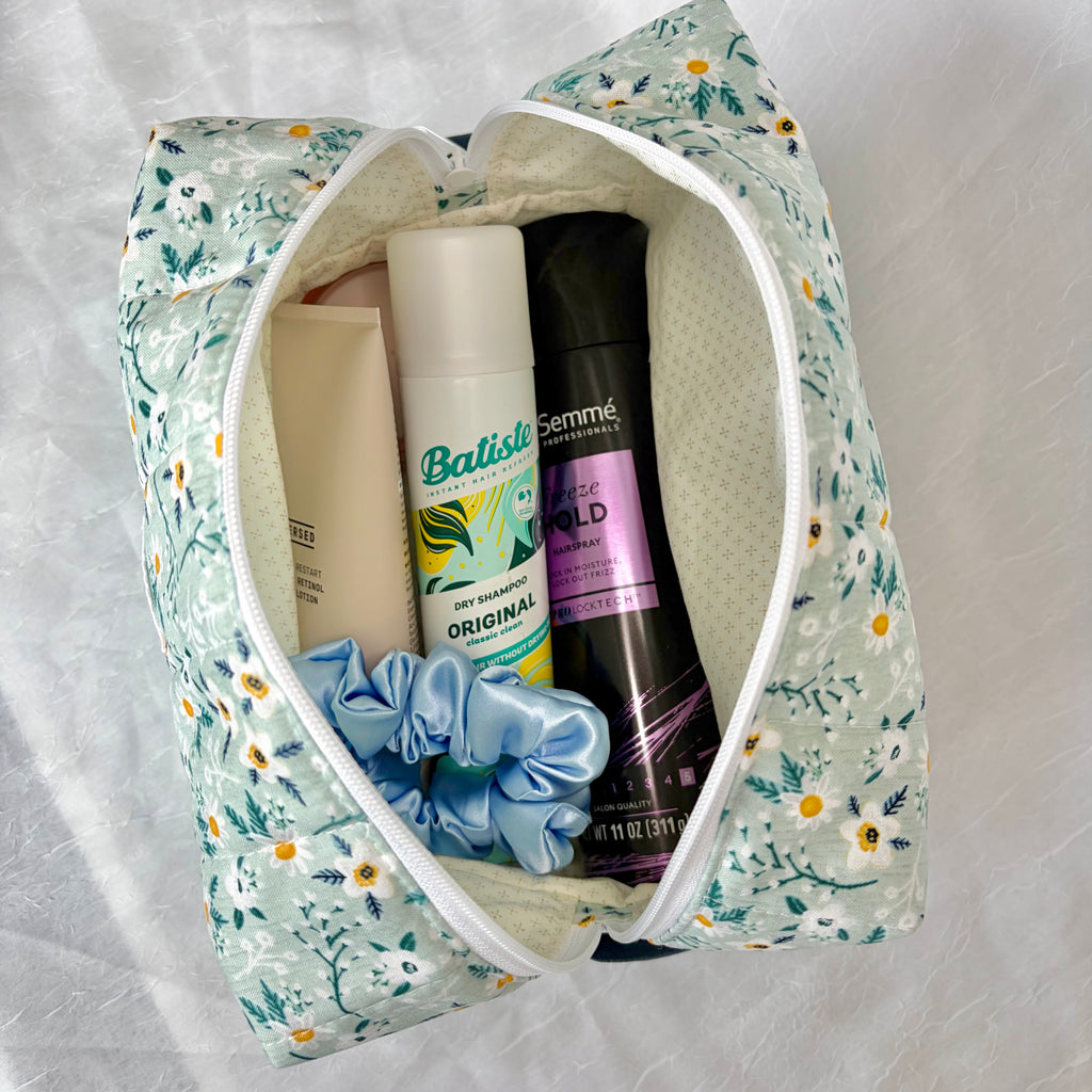A floral zippered travel or vanity pouch filled with toiletries, including dry shampoo, a light blue satin scrunchie, and other personal care items, laid on a white textured fabric background.