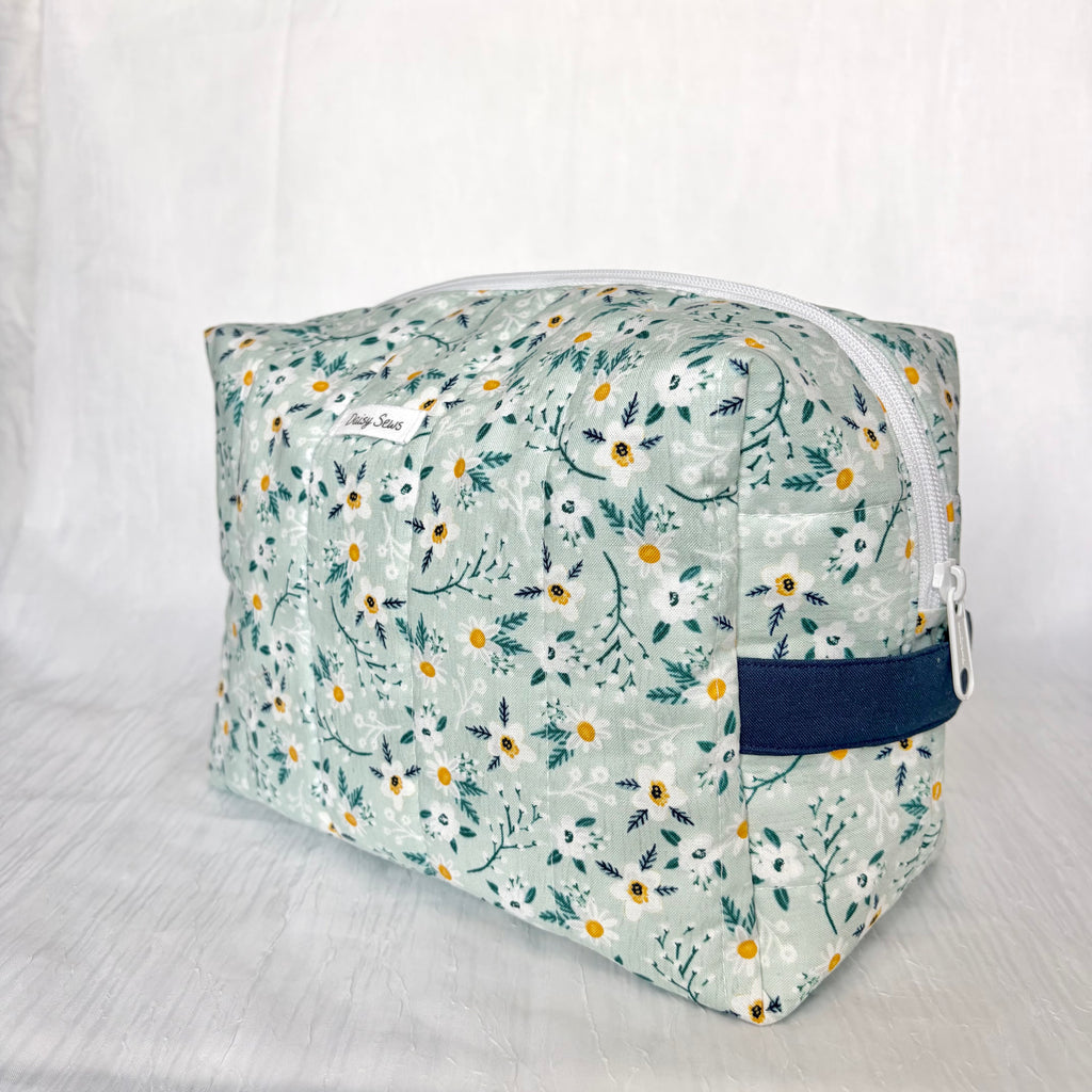 A rectangular zippered cosmetic pouch with a light sage green floral print, featuring white daisies with yellow centers, a navy blue handle and a white zipper.
