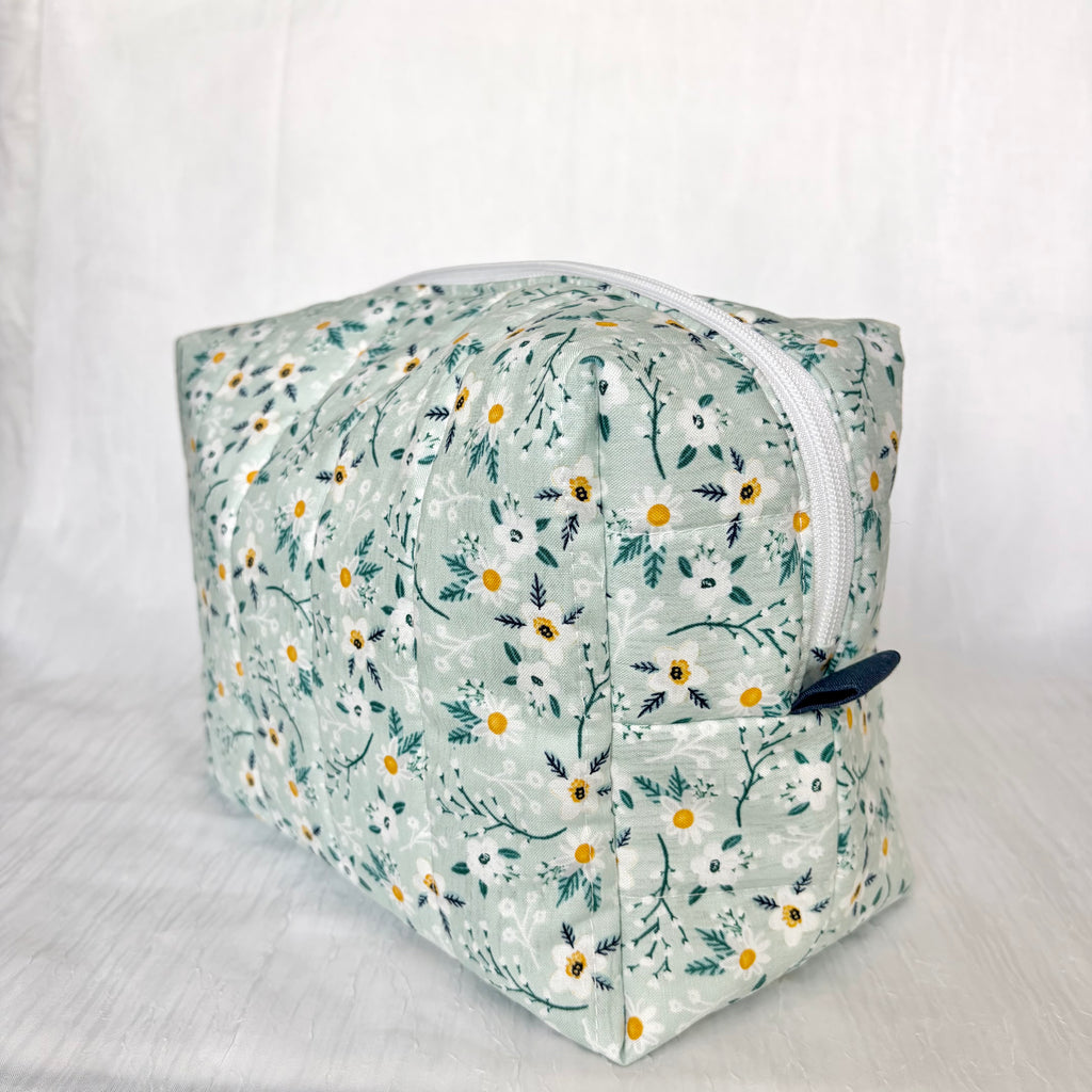 A quilted cosmetic pouch with a white floral print featuring daisies and green leaves on a pale mint background, shown on a white fabric backdrop.