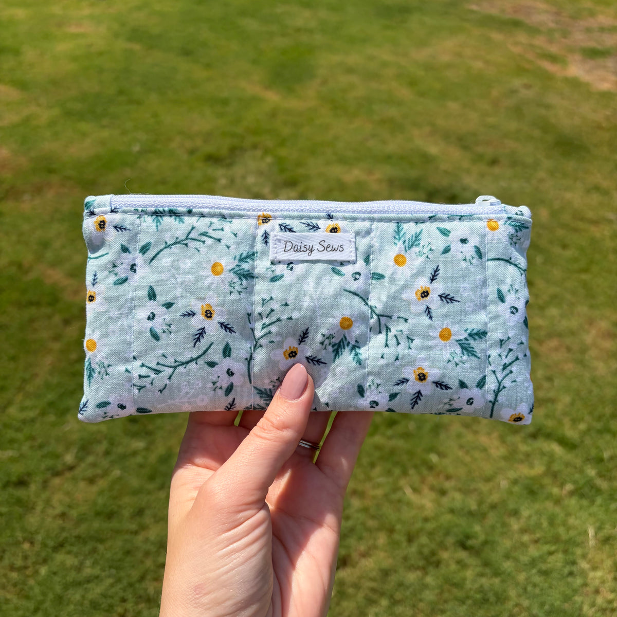 A hand holding a light blue quilted zipper pouch with a floral print of yellow daisies and green leaves, set against a blurred green grass background. The pouch features a white label that reads 'Daisy Sews'.
