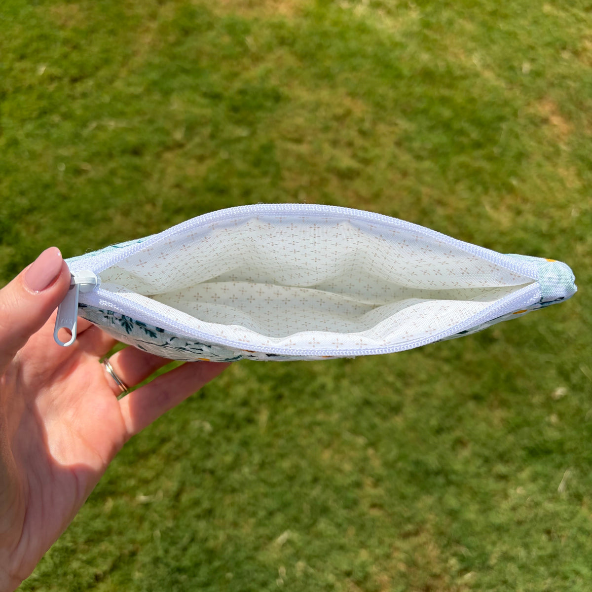 A hand holding an open zippered pouch outdoors on a grassy lawn. The pouch has a white interior with tiny blue polka dots and a light blue and white floral pattern on the exterior.