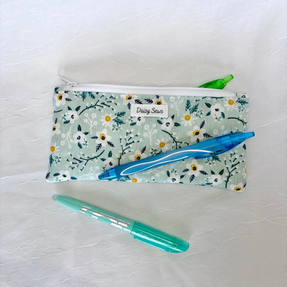 A handmade fabric zippered pencil case with a floral print in mint green, white, and yellow, featuring a white zipper and a small white label reading 'Daisy Sews'. A blue pen rests on top of the pouch, and a translucent mint green pen is placed below it on a textured white background.
