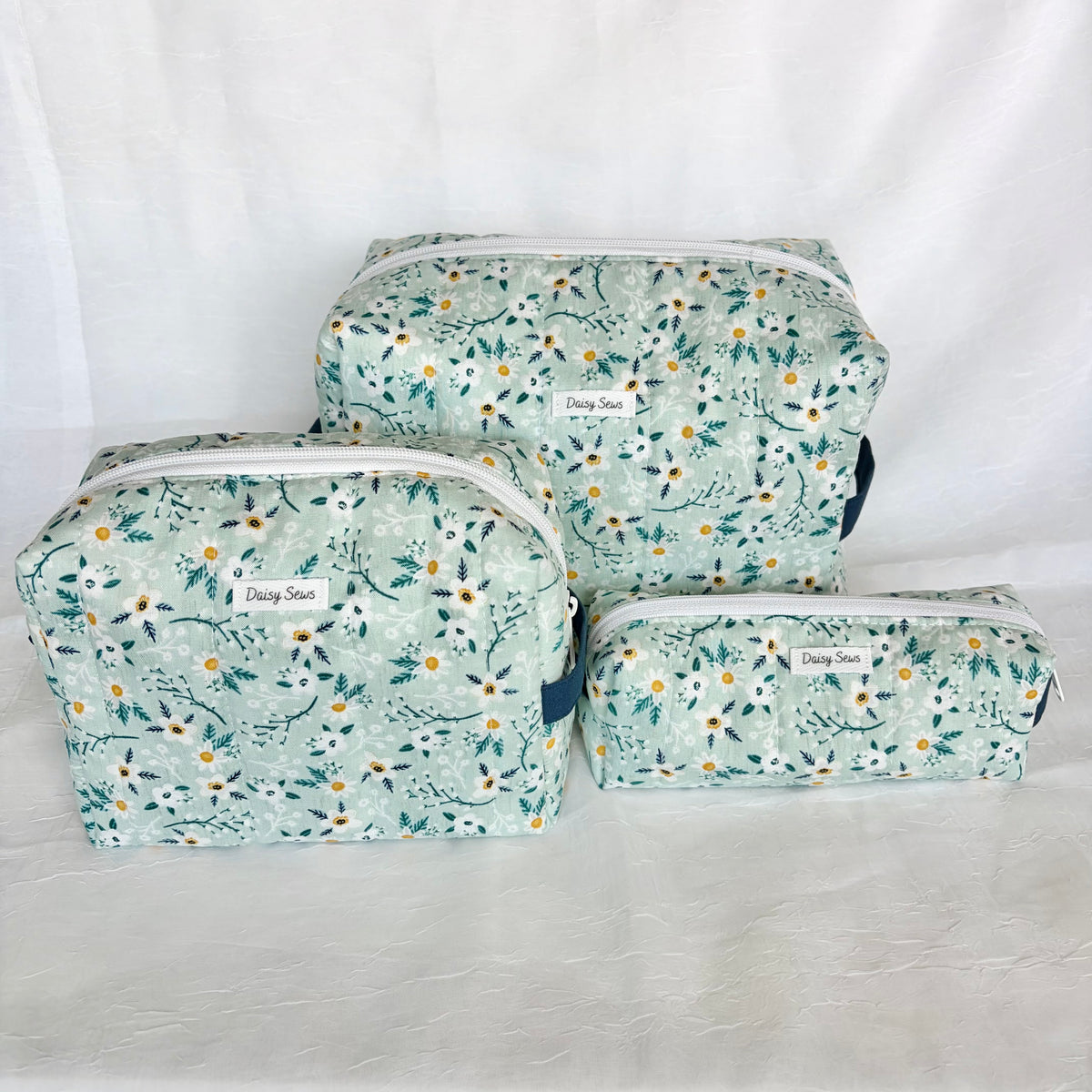 Three floral makeup bags in different sizes arranged on a white fabric backdrop. The bags feature a light blue green background with a delicate white daisy and green leaf pattern, along with white zippers and small labels reading 'Daisy Sews'.