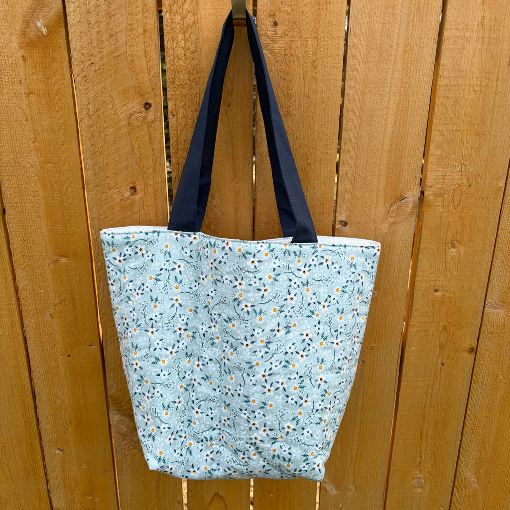 Back side of blue tote bag with white flowers and navy handles handing on a wooden fence