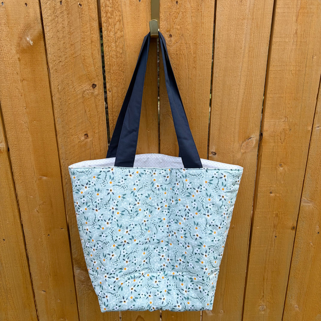 white Floral on blue tote bag with navy handles hanging on a wooden fence.