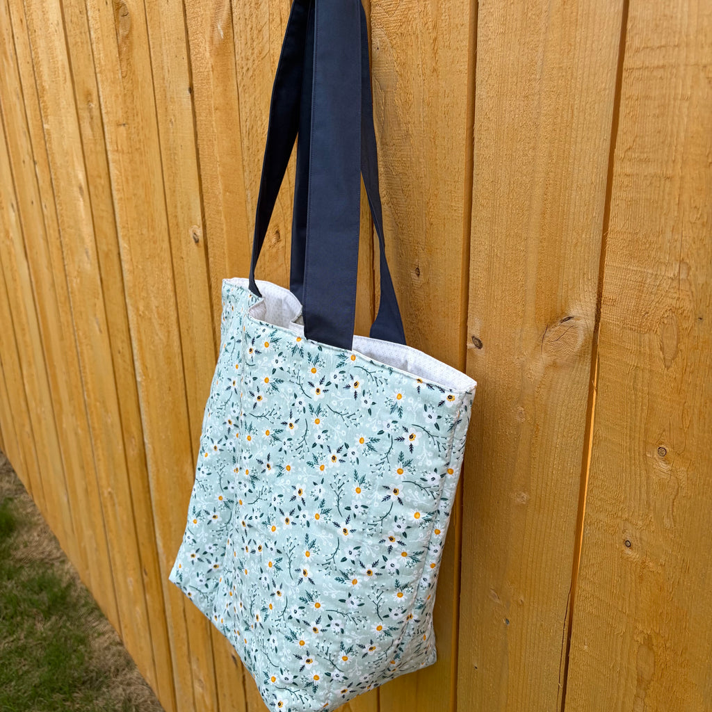  Side view of blue and white floral-patterned tote bag with navy straps against a wooden fence.