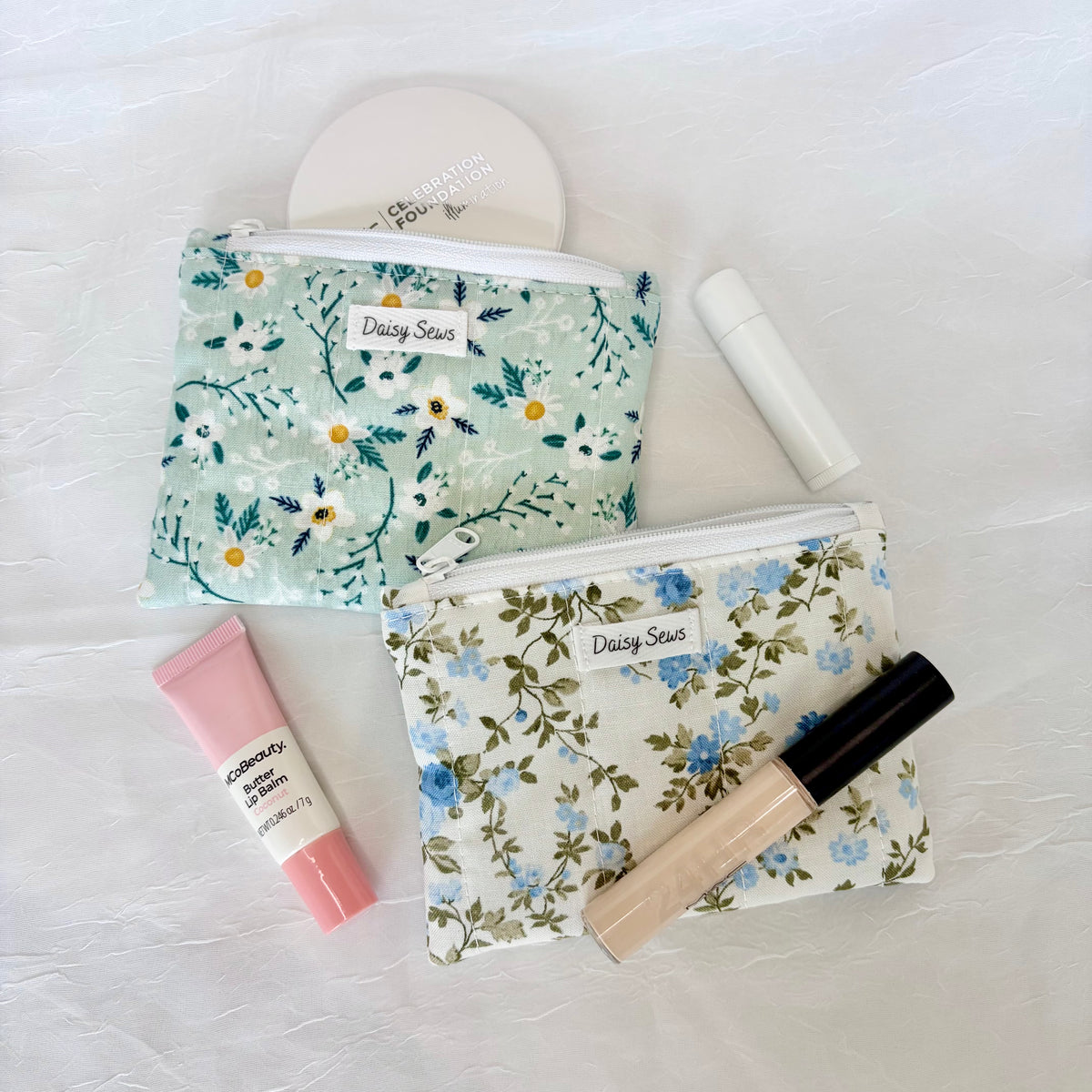 Two floral cosmetic pouches from Daisy Seis are arranged on a white textured fabric background with various makeup items. The top pouch is light green with white and yellow flowers, and the bottom pouch is white with blue and green flowers.