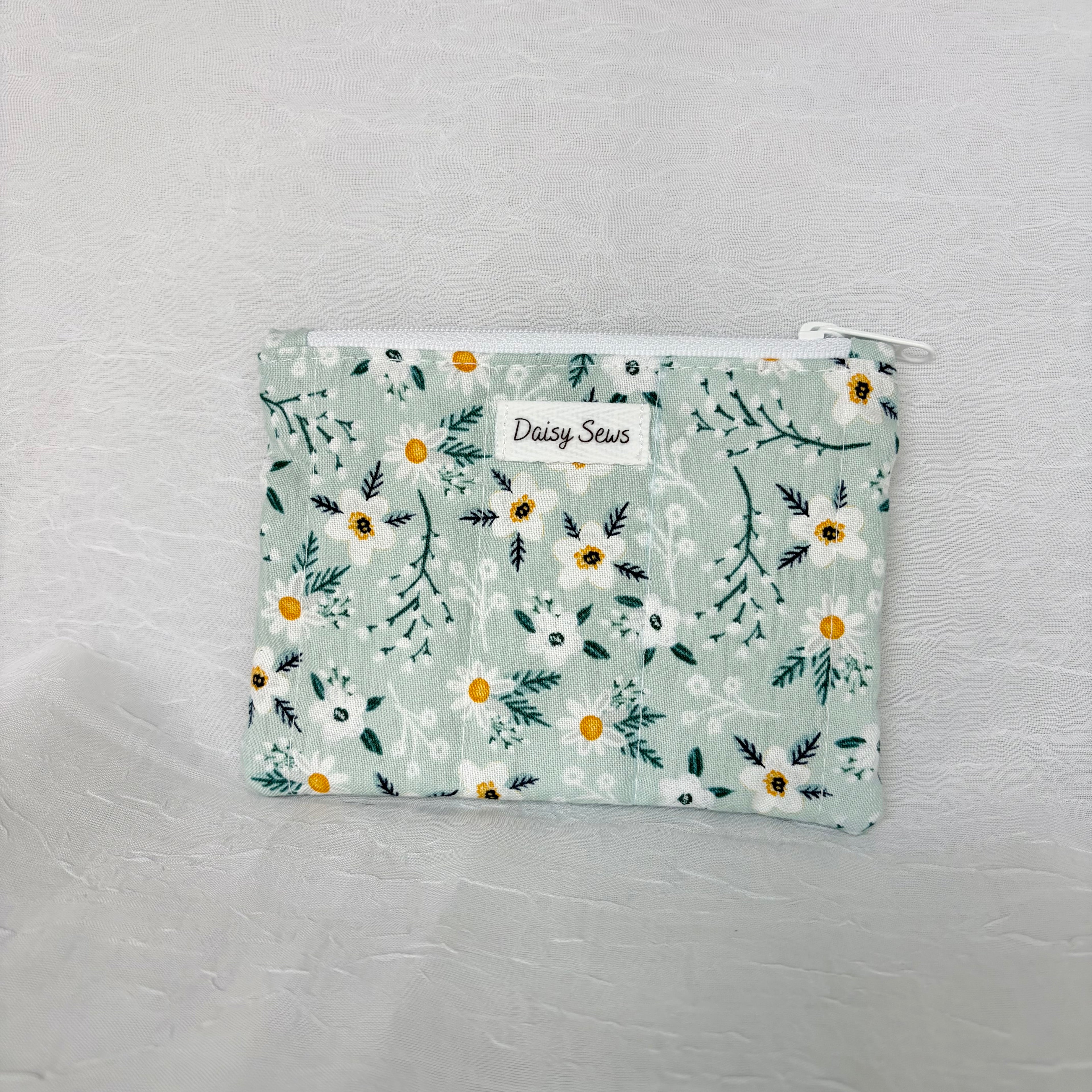 Blue fabric with whit flowers and teal leaf patterned small pouch with white zipper on white background