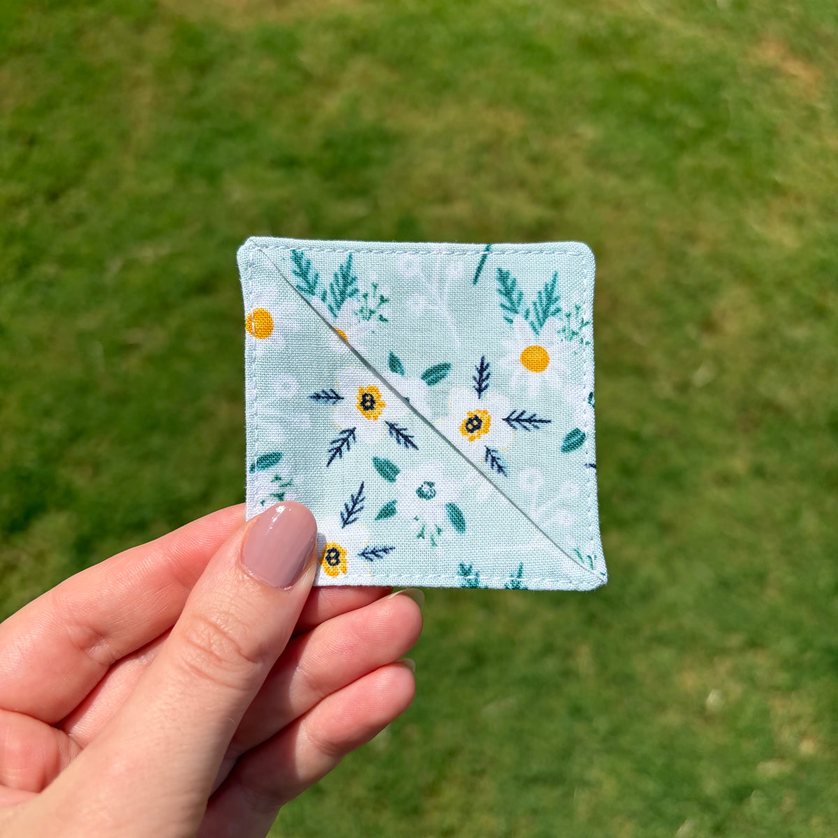 A hand with pink nail polish holds a square, diamond-shaped fabric patch featuring a light blue and white floral pattern with green leaves and small yellow flowers, set against a blurred green grass background.