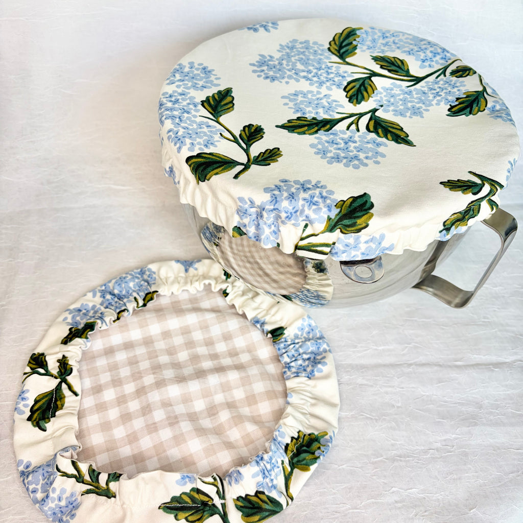 Blue hydrangea with green leaves on white fabric bowl cover displayed on a metal bowl with a handle and a matching cover with beige gingham interior on a light gray background