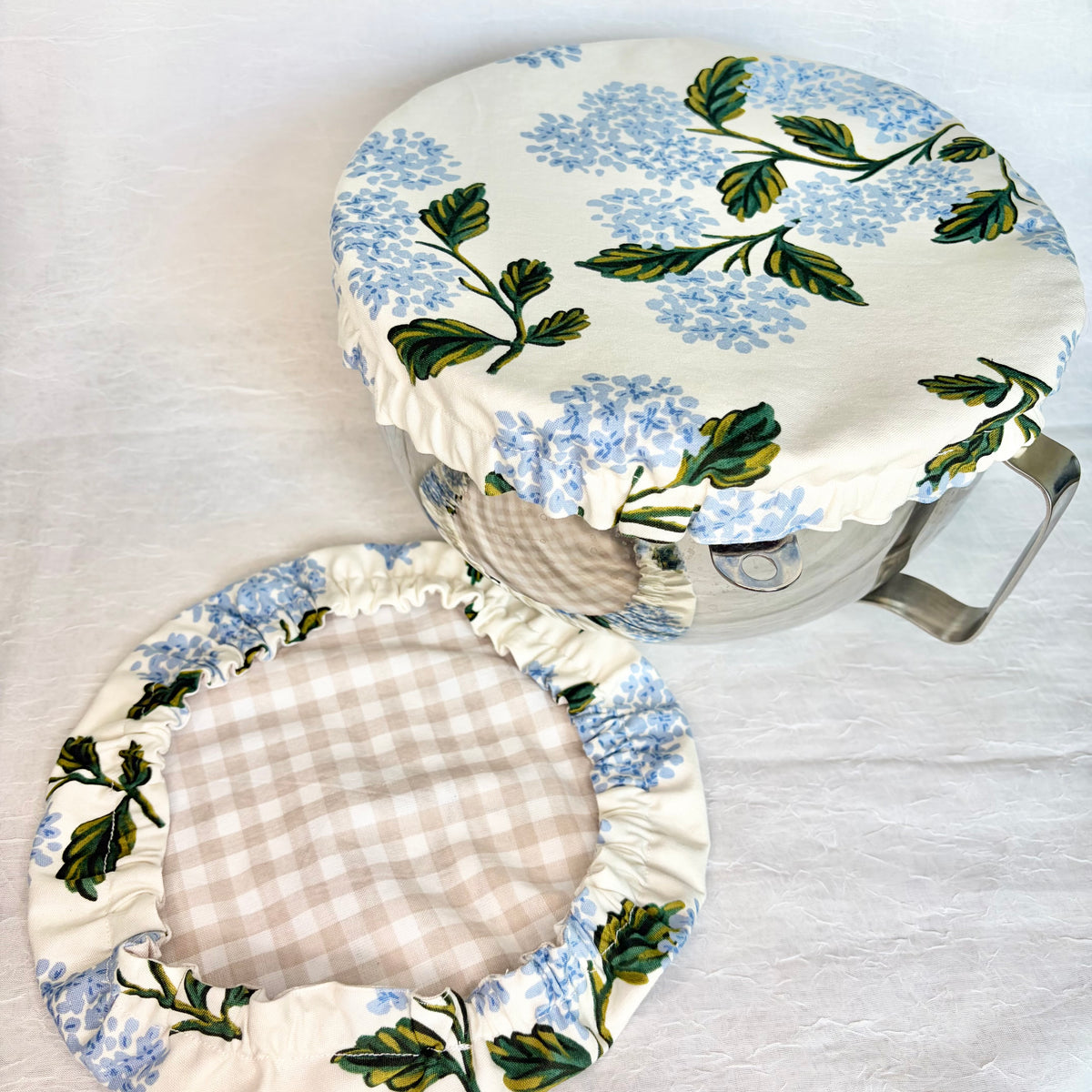 Blue hydrangea with green leaves on white fabric bowl cover displayed on a metal bowl with a handle and a matching cover with beige gingham interior on a light gray background