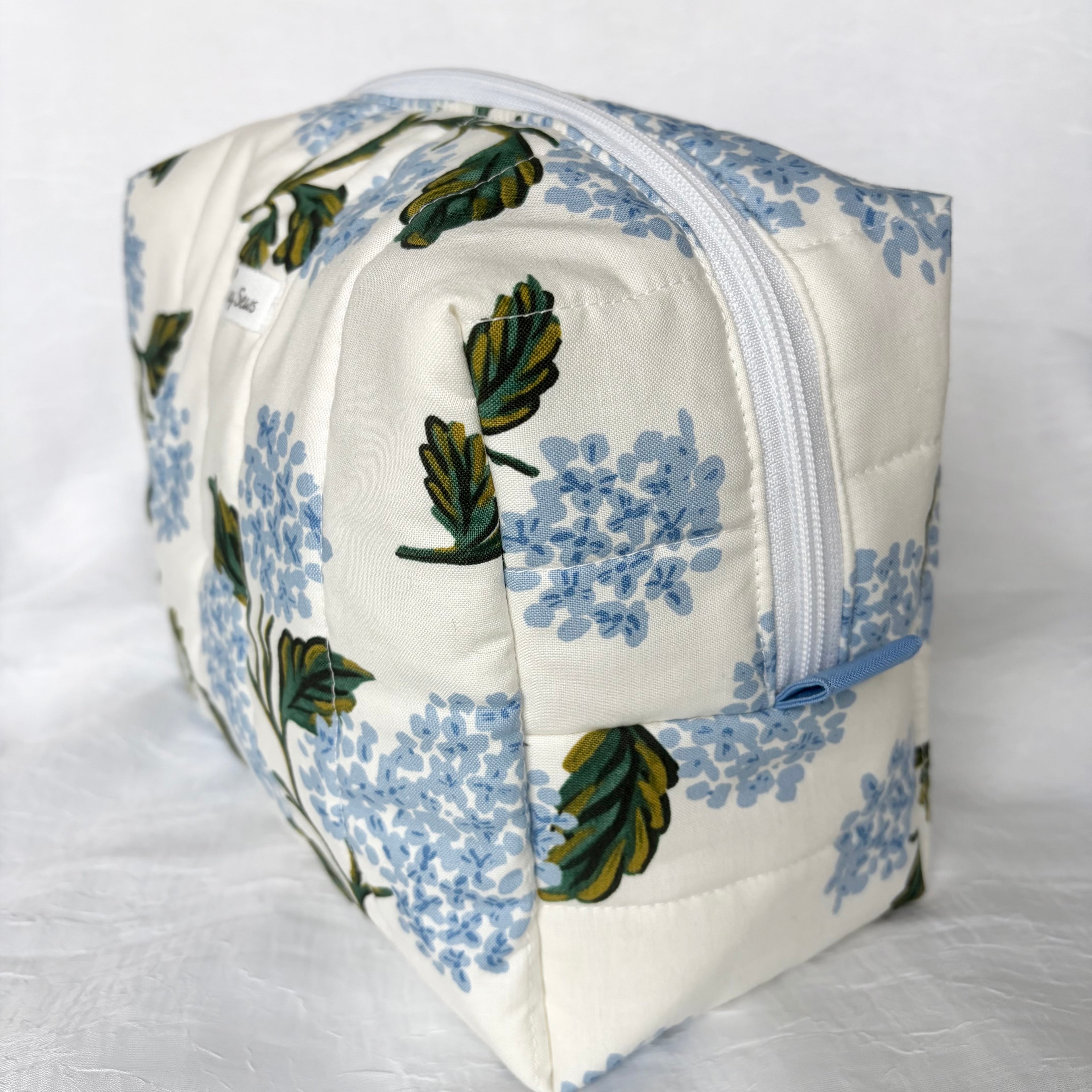 Side view of blue Pull Tab and white zipper on large blue hydrangea and green leaf designed boxy bag with white label on white background
