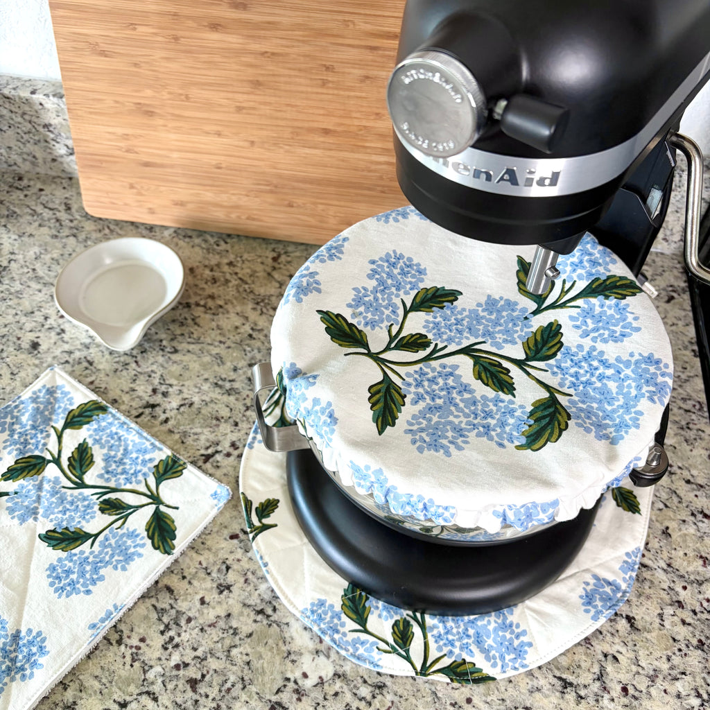 A KitchenAid stand mixer on a granite countertop with a blue and green floral print fabric cover, alongside a matching cloth napkin and a small white bowl.