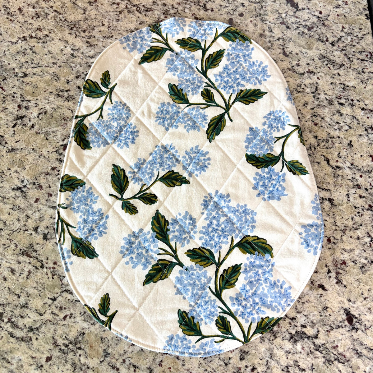 An oval quilted fabric mat, featuring a blue hydrangea floral pattern on a cream background, laid flat on a speckled granite countertop. Bowl Lift Size