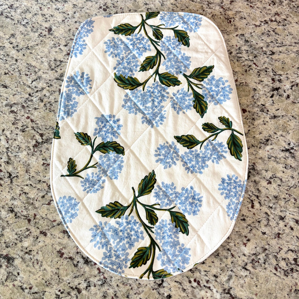 An oval quilted fabric mat, featuring a light blue hydrangea floral print with green leaves on an off-white background, laid flat on a speckled granite countertop. Head Tilt Size
