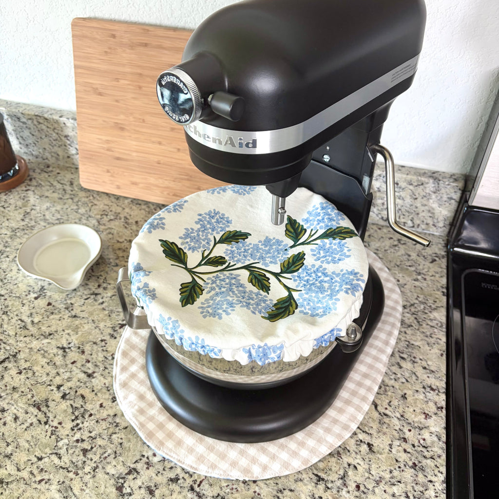 A black KitchenAid stand mixer sits on a granite kitchen countertop, covered with a white fabric dust cover printed with blue flowers and green leaves.