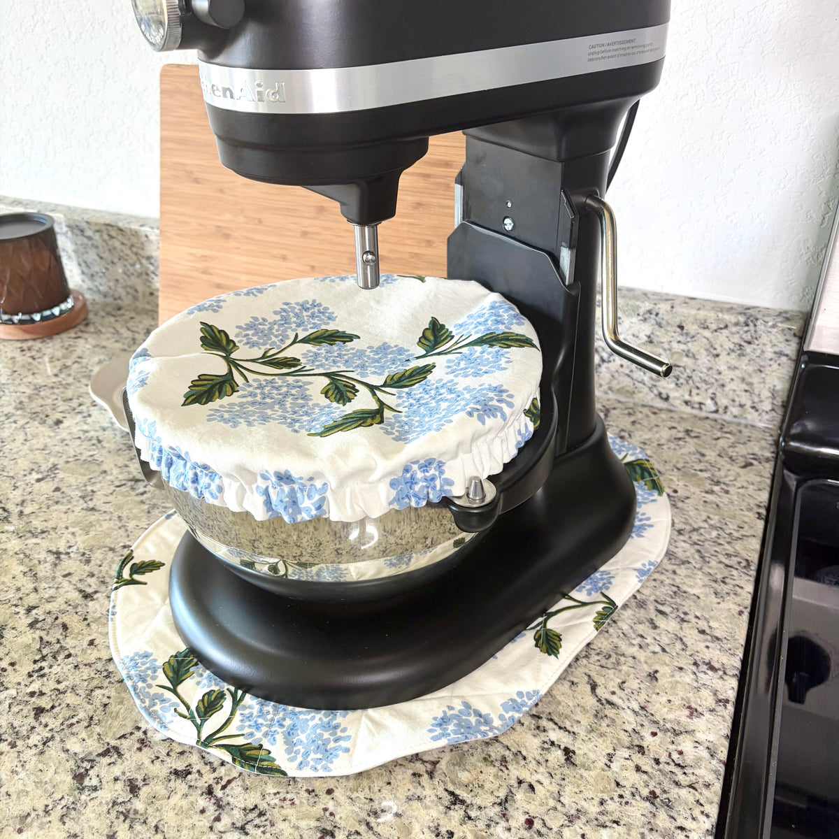 A black stand mixer with a glass bowl covered by a blue and green floral fabric dust cover, sitting on a granite kitchen countertop.