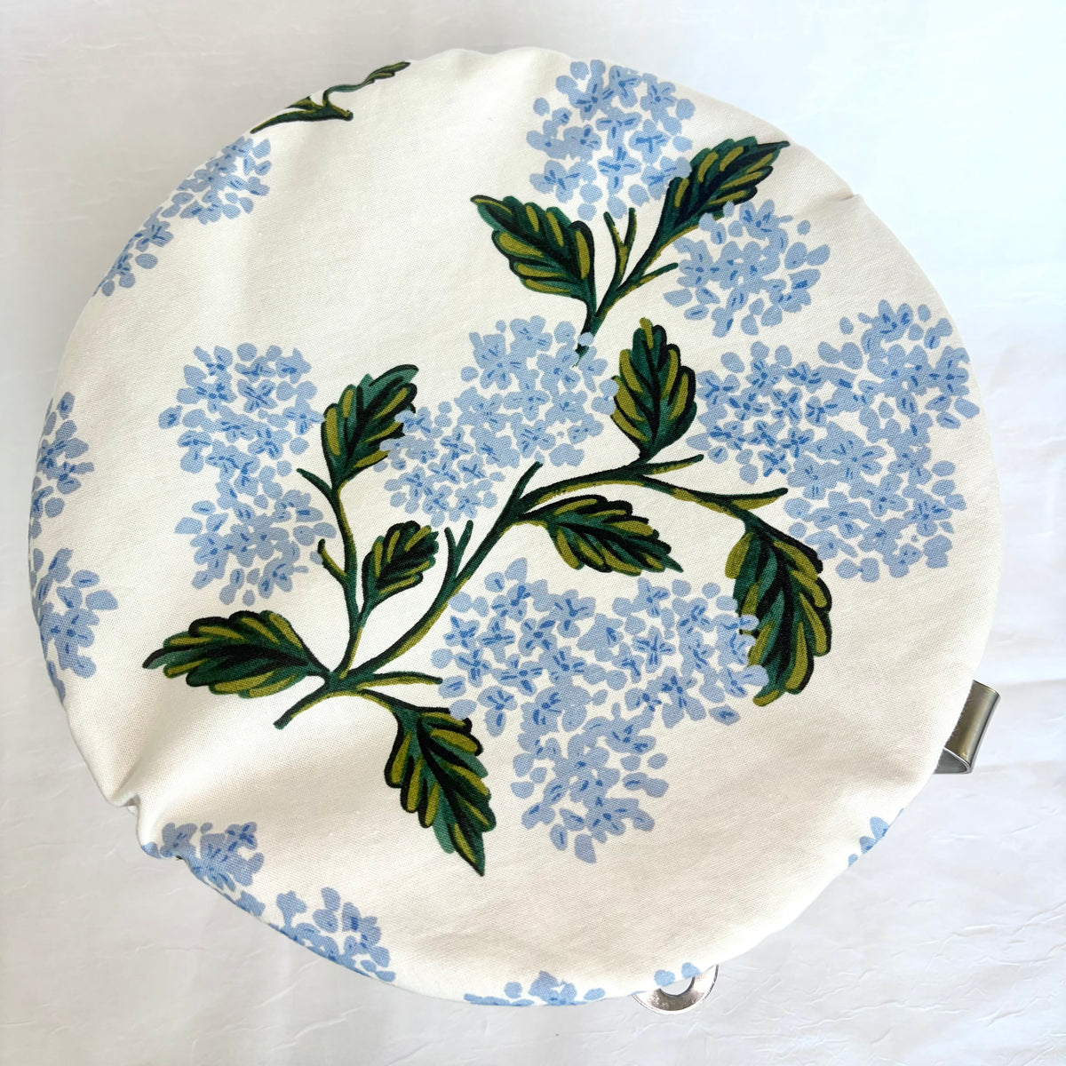 A round fabric-covered bowl with a cream background, featuring a printed pattern of light blue hydrangea flowers and green leaves. A small metal handle is visible on the right side.