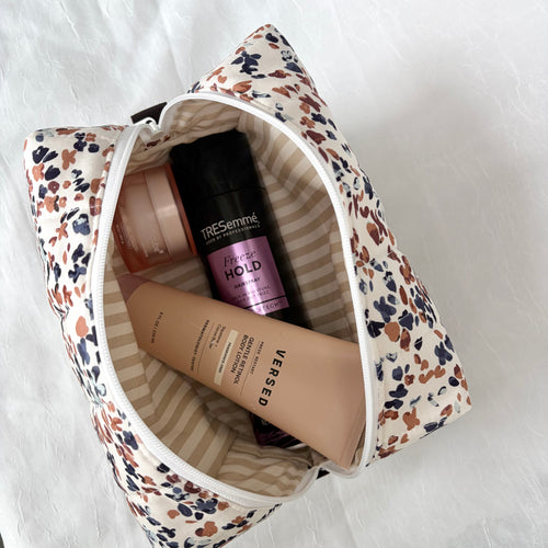 tan lotion bottle black hairspray bottle, and pink circular container inside a toiletry bag with  tan striped interior and blue and mauve speckles on the outside.