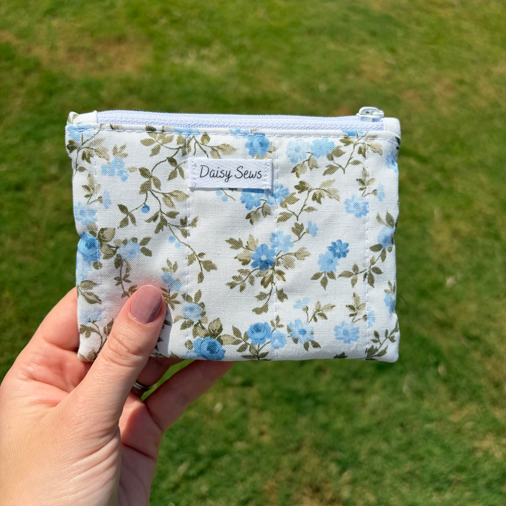 A hand holds a small zippered pouch with a white background and a blue floral print. The pouch has a white label that reads 'Daisy Sews' and is photographed outdoors against a blurred green grass background.