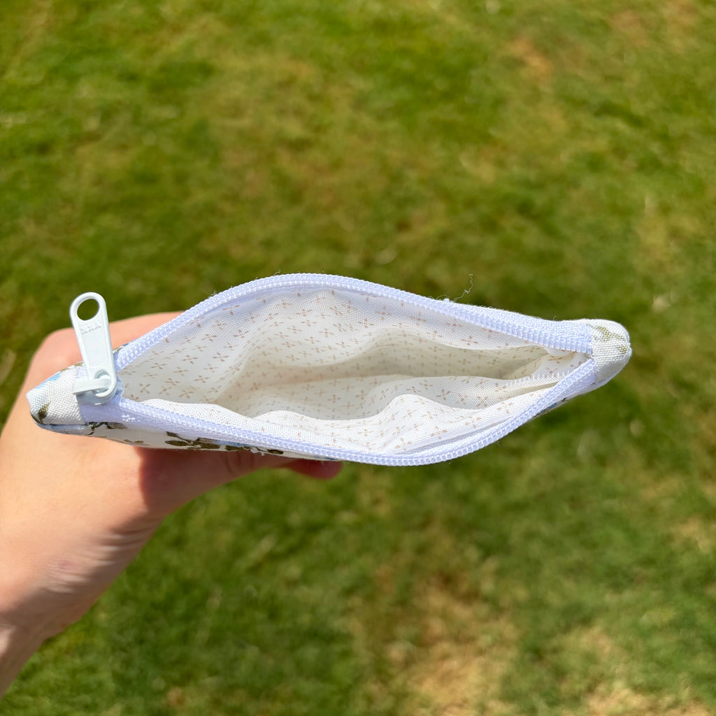 A hand holding an open small zip pouch outdoors against a blurred green grass background. The pouch has a white zipper, a white patterned lining, and a white pull tab.