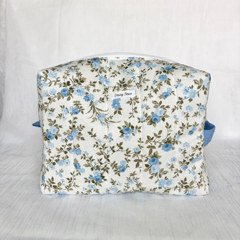 A fabric cosmetic bag with a blue and green floral pattern on a white background, featuring a small white label that reads 'Daisy Sews' and a blue strap on the sides, set against a white fabric backdrop.