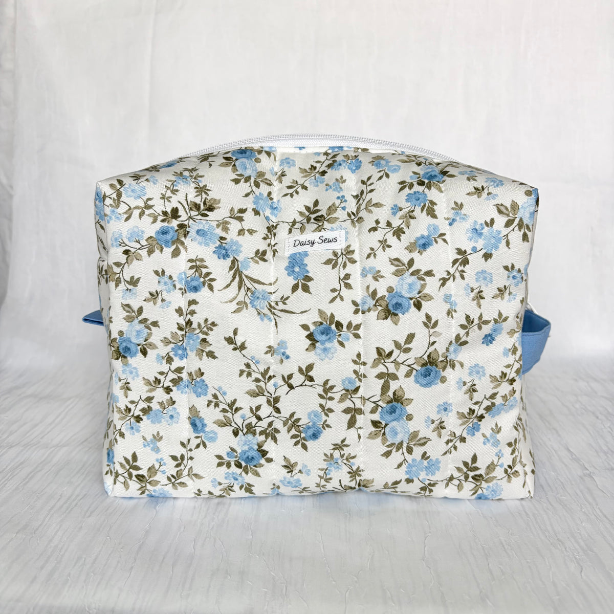A fabric cosmetic bag with a blue and green floral pattern on a white background, featuring a small white label that reads 'Daisy Sews' and a blue strap on the sides, set against a white fabric backdrop.