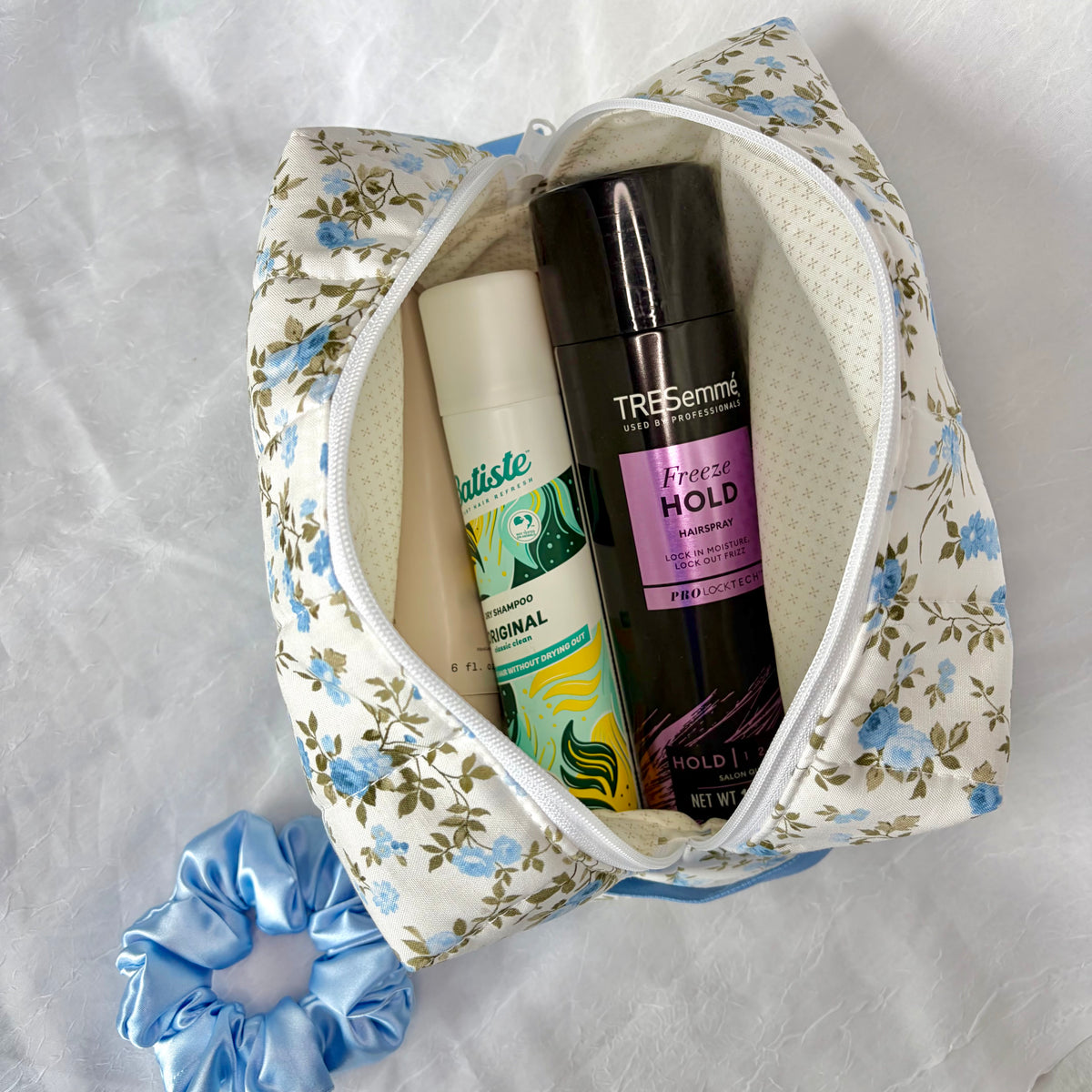 A floral zippered cosmetic pouch filled with toiletries, including hairspray and dry shampoo on a white fabric background with a blue satin scrunchie.
