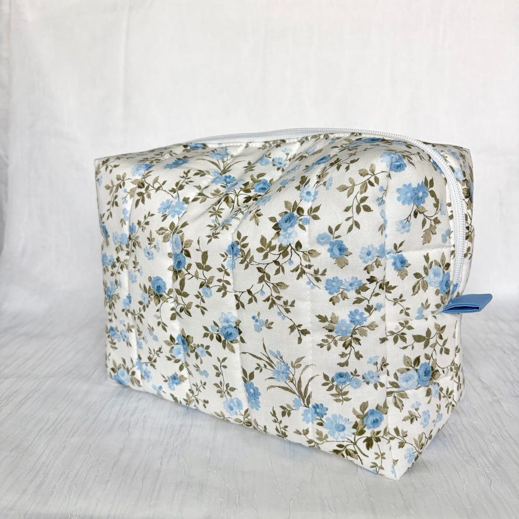 A rectangular zippered cosmetic bag with a white background and a blue and green floral pattern, shown on a white textured fabric backdrop.