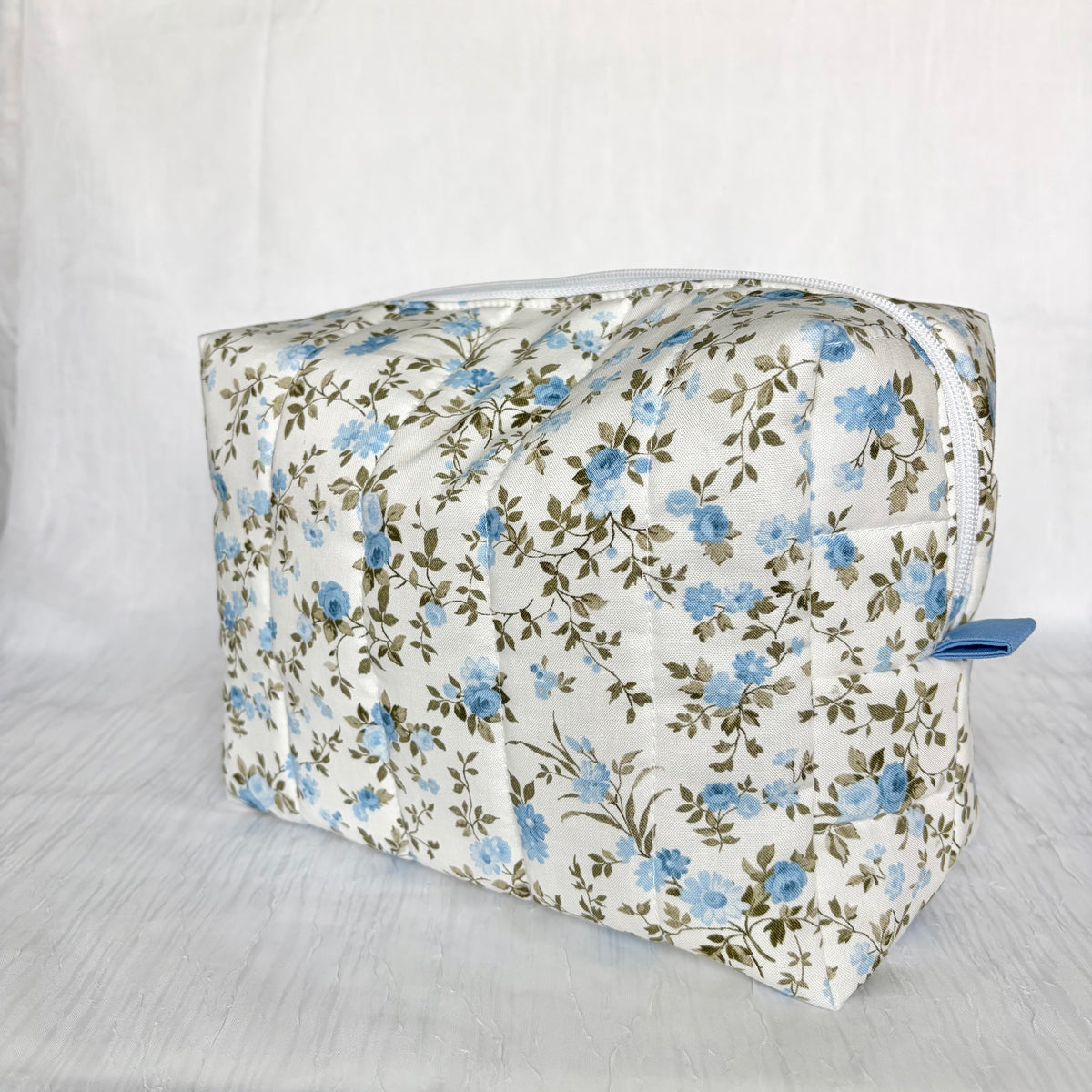 A rectangular zippered cosmetic bag with a white background and a blue and green floral pattern, shown on a white textured fabric backdrop.