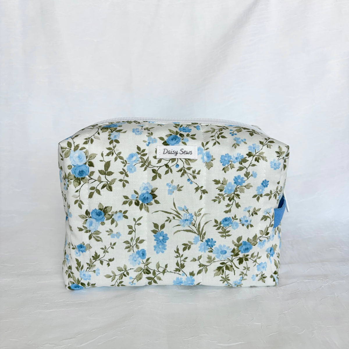 A fabric zippered cosmetic pouch with a blue floral print on a cream background, featuring a small white label that reads 'Daily Sews'.