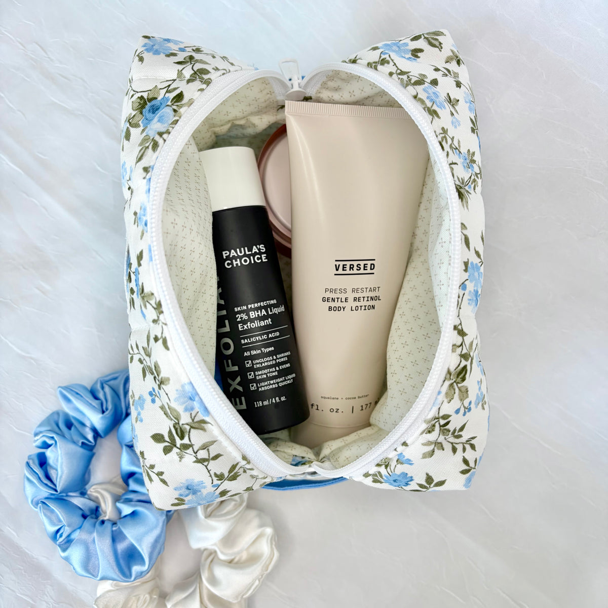 A floral fabric cosmetic pouch with a blue and green print, shown open with various toiletries inside, including a black bottle labeled 'PAULA'S CHOICE' and a beige tube labeled 'VERSED'. A light blue satin scrunchie and a white satin ribbon are placed beside the pouch on a white textured fabric background.