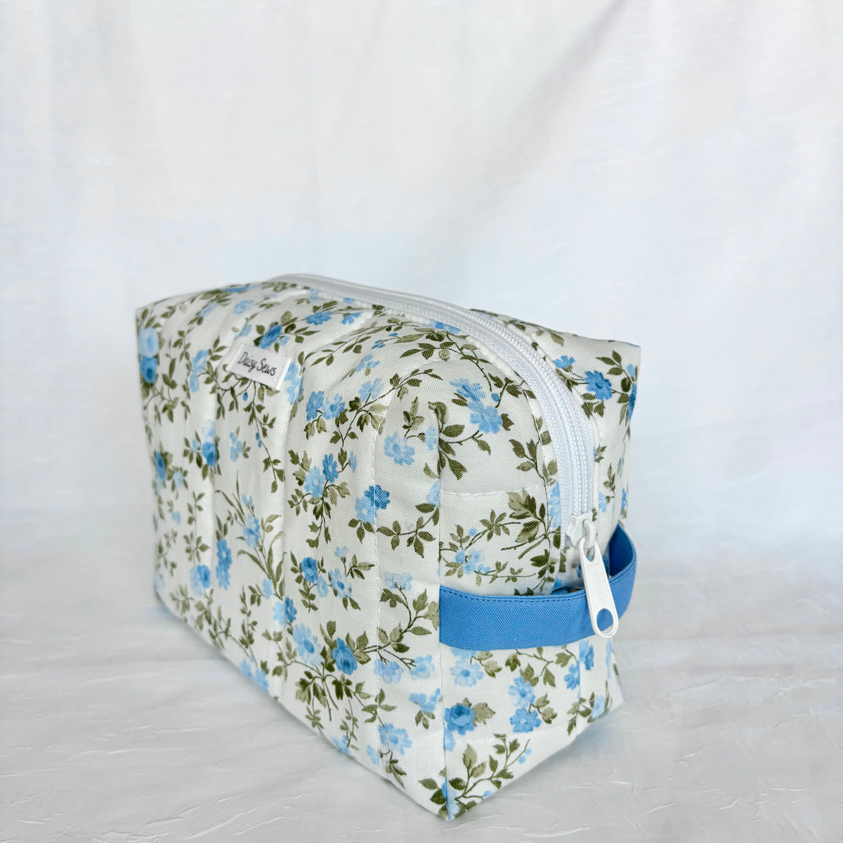 A rectangular zippered cosmetic pouch with a blue and green floral print on a cream background, featuring a blue handle and a white zipper.