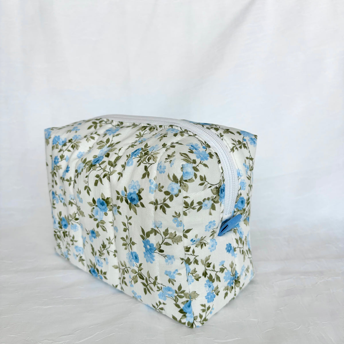 A rectangular zippered pouch with a blue and green floral pattern on a white background, photographed against a white fabric backdrop.