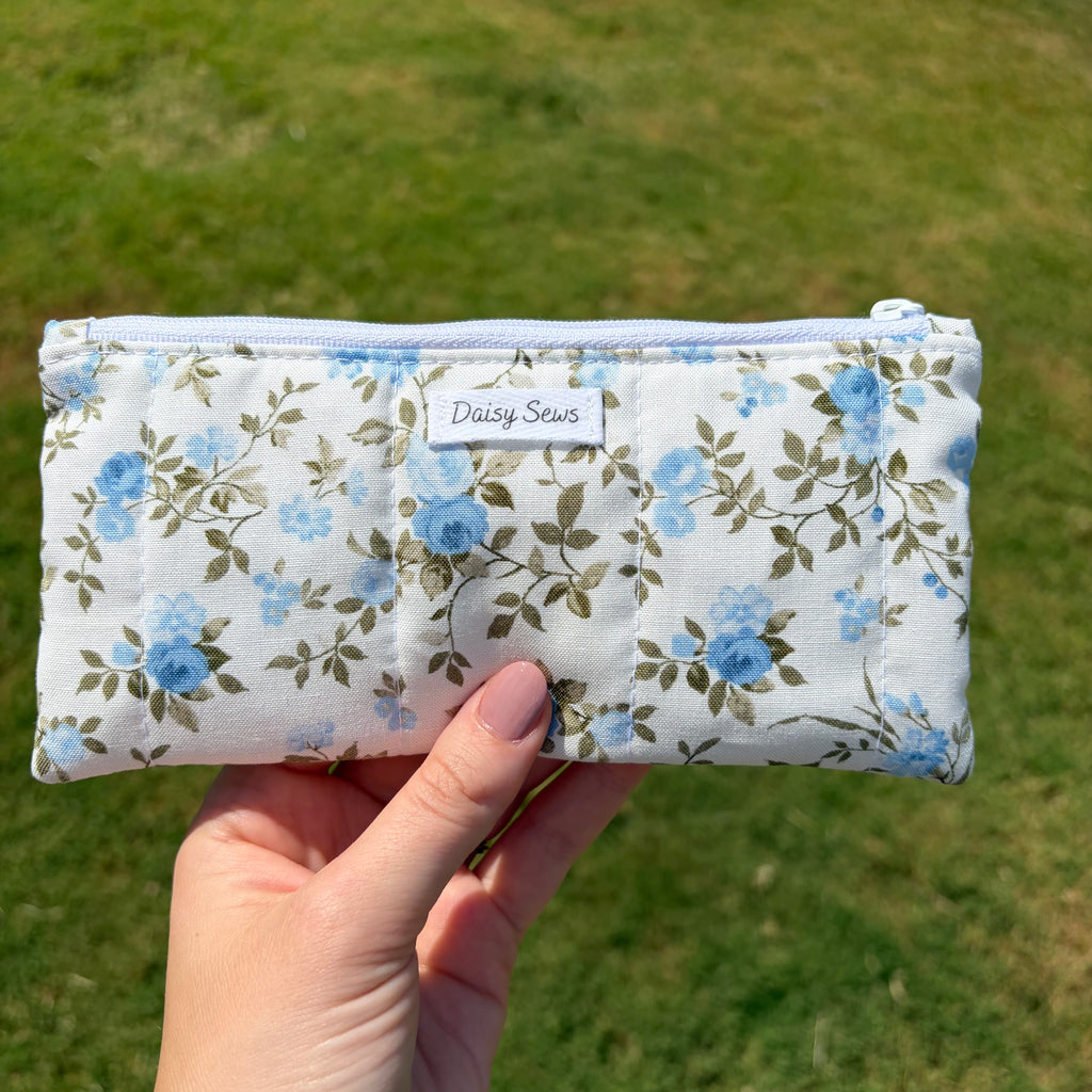 A hand holding a quilted fabric pouch with a blue floral print, set against a blurred green grass background. The pouch features a white label that reads 'Daisy Sews'.