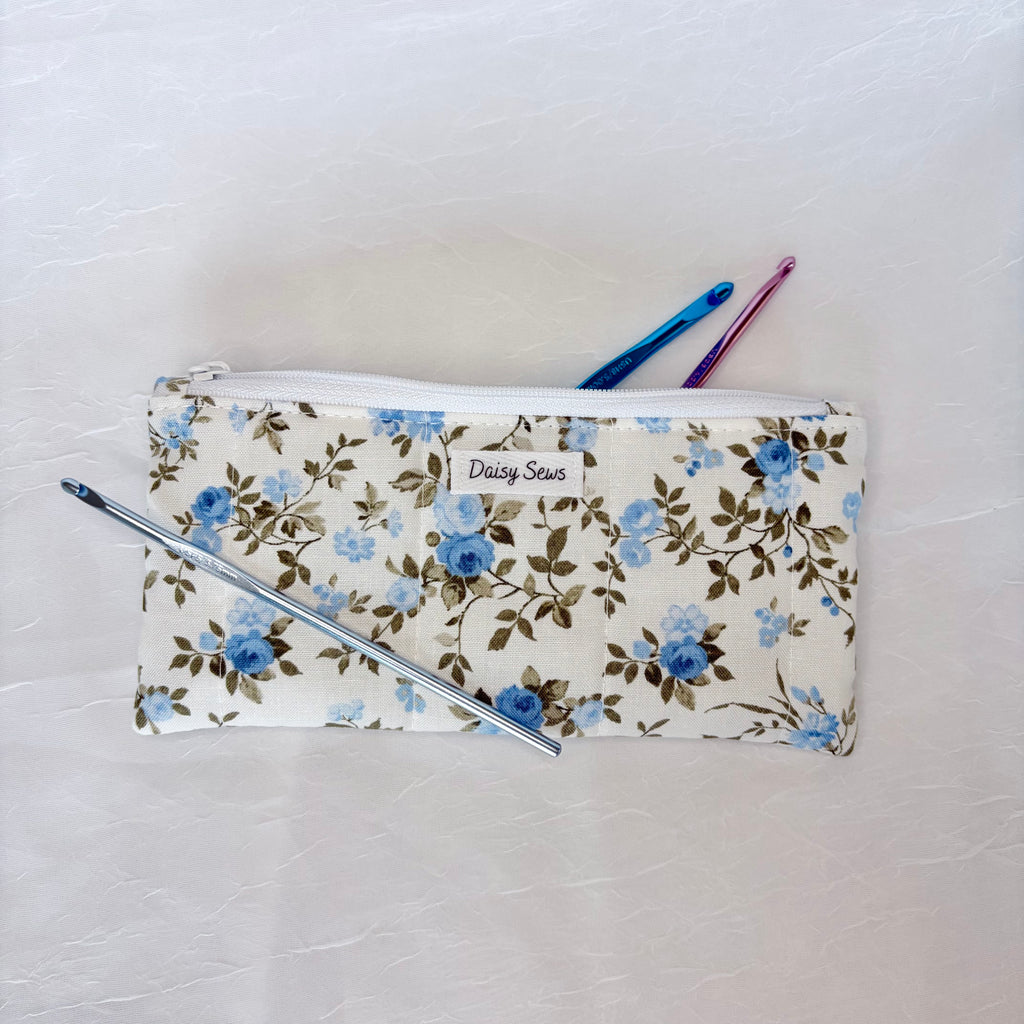 A fabric zippered pouch with a blue and brown floral pattern on a cream background, featuring a white zipper and a small 'Daisy Sews' label. A silver pen and two crochet hooks are placed on and around the pouch, all on a light textured surface.