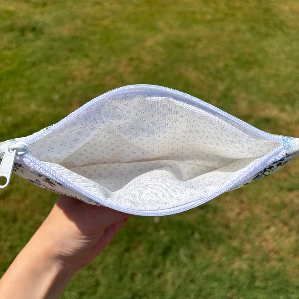 A hand holding open a zippered pouch or small bag outdoors, showing the white interior with a tiny dotted pattern and a light blue zipper. The background is blurred green grass.