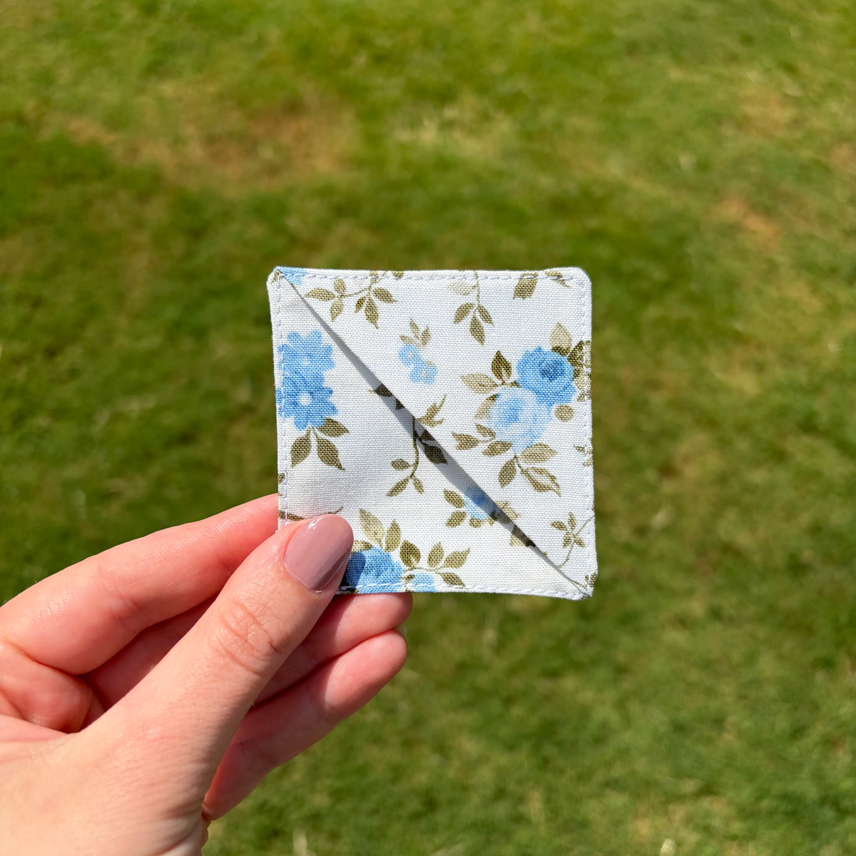 A hand holding a small square fabric item with a blue floral print on a white background, photographed outdoors against a blurred green grass background.
