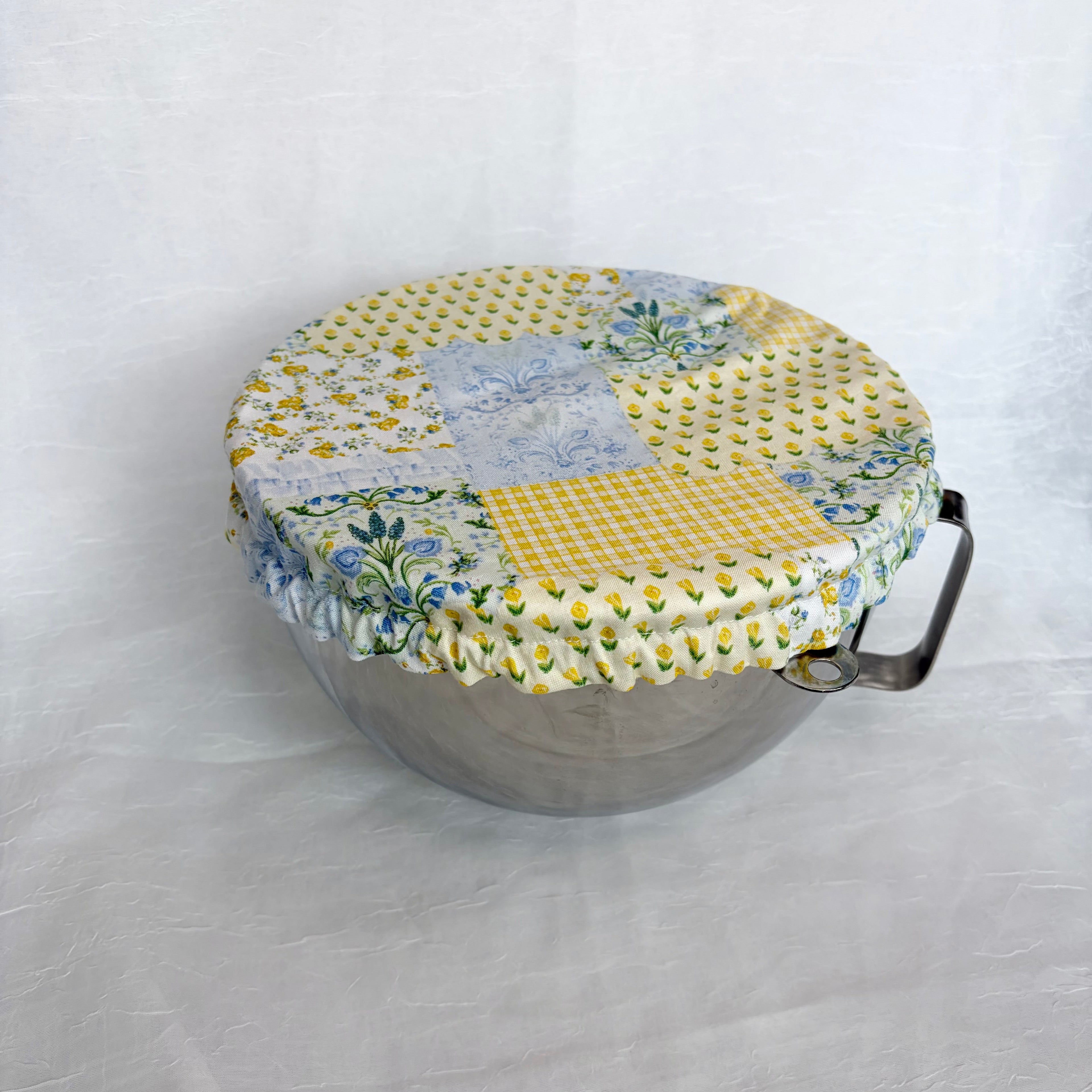 Blue and yellow floral patchwork cover on a silver metal bowl with handle on white background