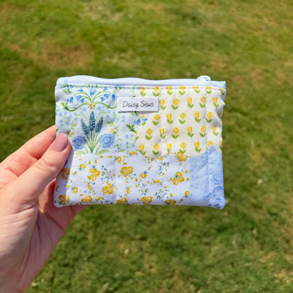 A hand holds a small zippered pouch with a light blue and white floral patchwork print, featuring yellow daisies, blue flowers, and a 'Daisy Sews' brand label, against a blurred green grass background.