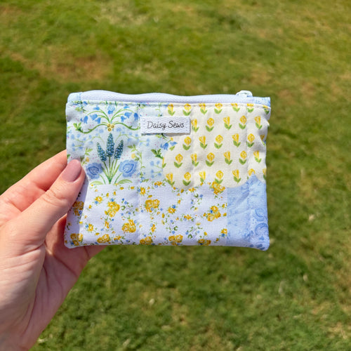 A hand holds a small zippered pouch with a light blue and white floral patchwork print, featuring yellow daisies, blue flowers, and a 'Daisy Sews' brand label, against a blurred green grass background.