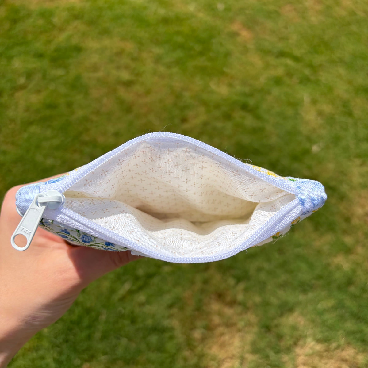 A hand holding an open fabric zip pouch with a light blue floral pattern and white interior, set against a blurred green grass background.
