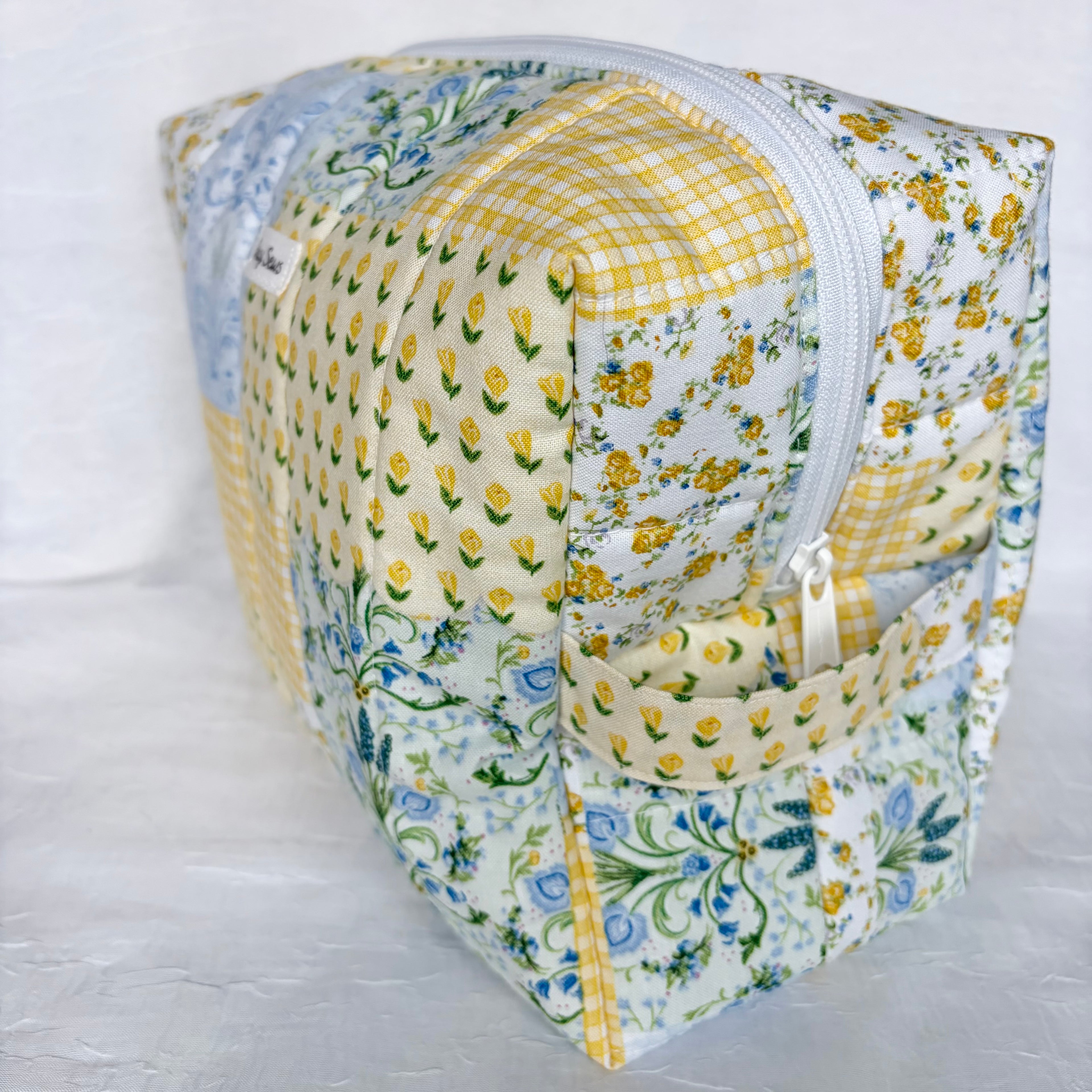 side view of handle and white zipper on blue and yellow floral patchwork boxy bag with white label against white background