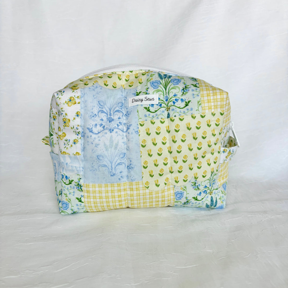 A square patchwork cosmetic pouch with a top zipper, featuring yellow tulip print, blue damask pattern, and gingham checks, with a 'Daisy Sews' label on the front.