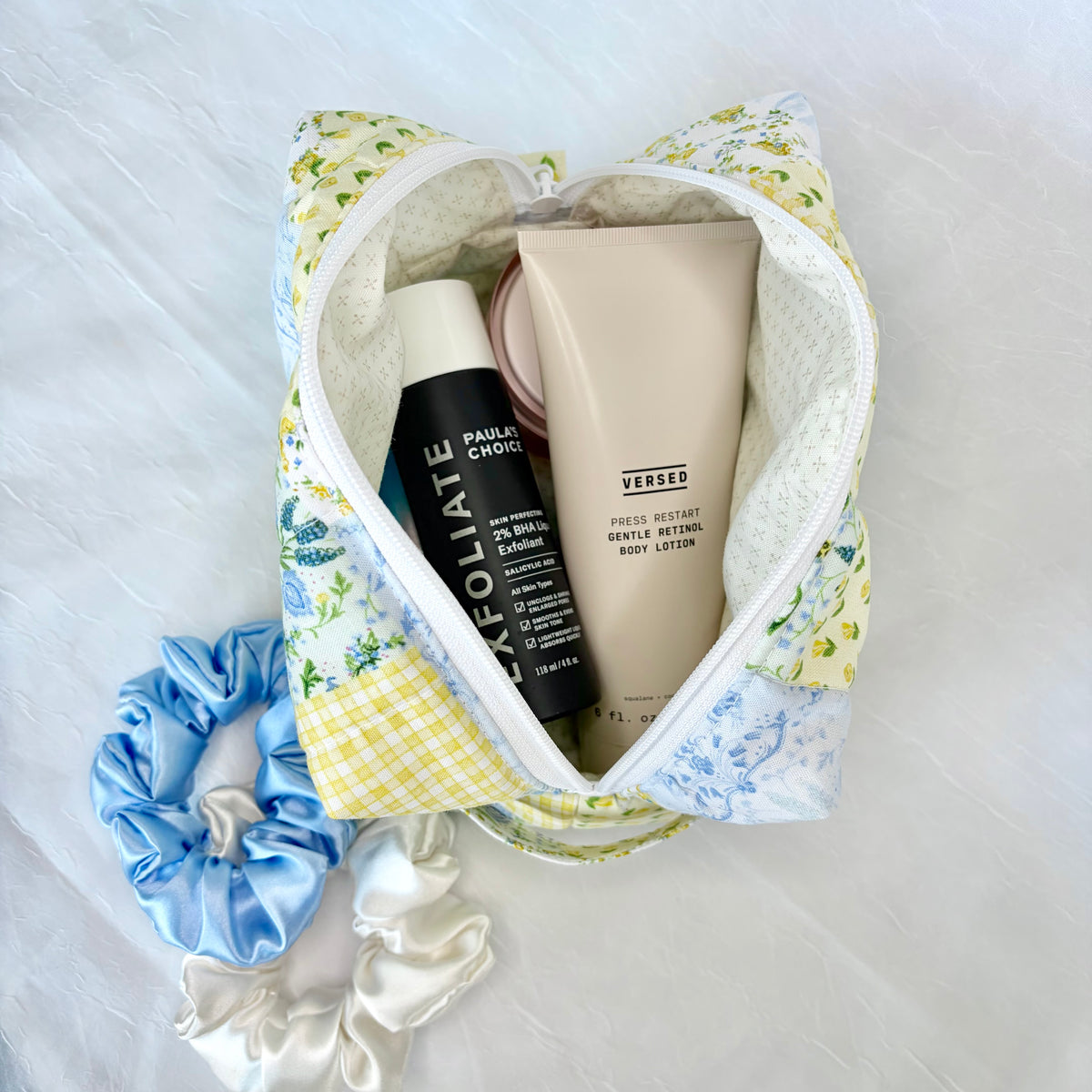 A floral and gingham patchwork cosmetic pouch filled with skincare items, including a black bottle labeled 'Paulas Choice!' and a beige tube labeled 'VERSED', styled on a white fabric background with blue and white satin scrunchies.