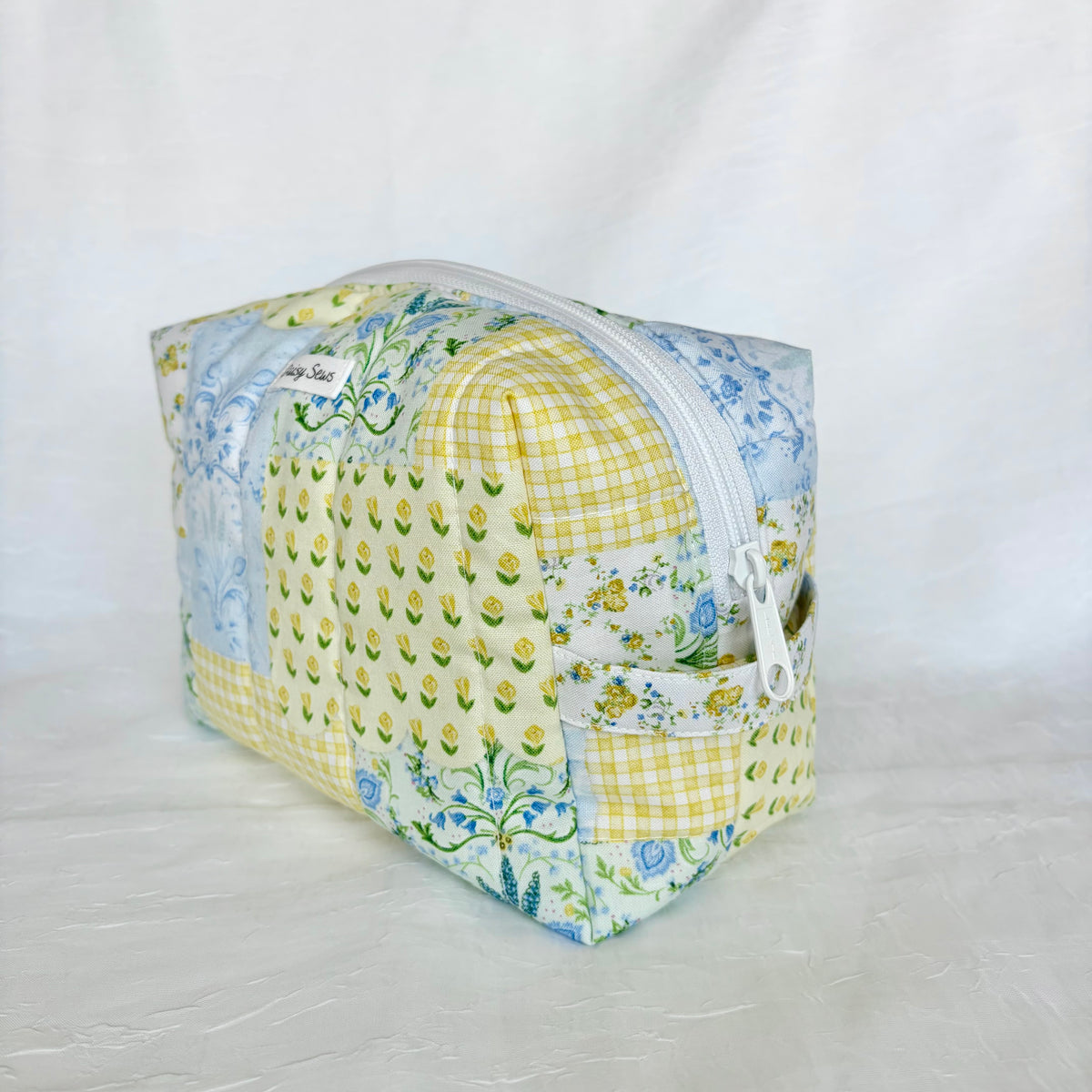 A square zippered pouch made from a patchwork of yellow gingham, yellow tulip print, and blue floral fabrics, set against a white fabric backdrop.