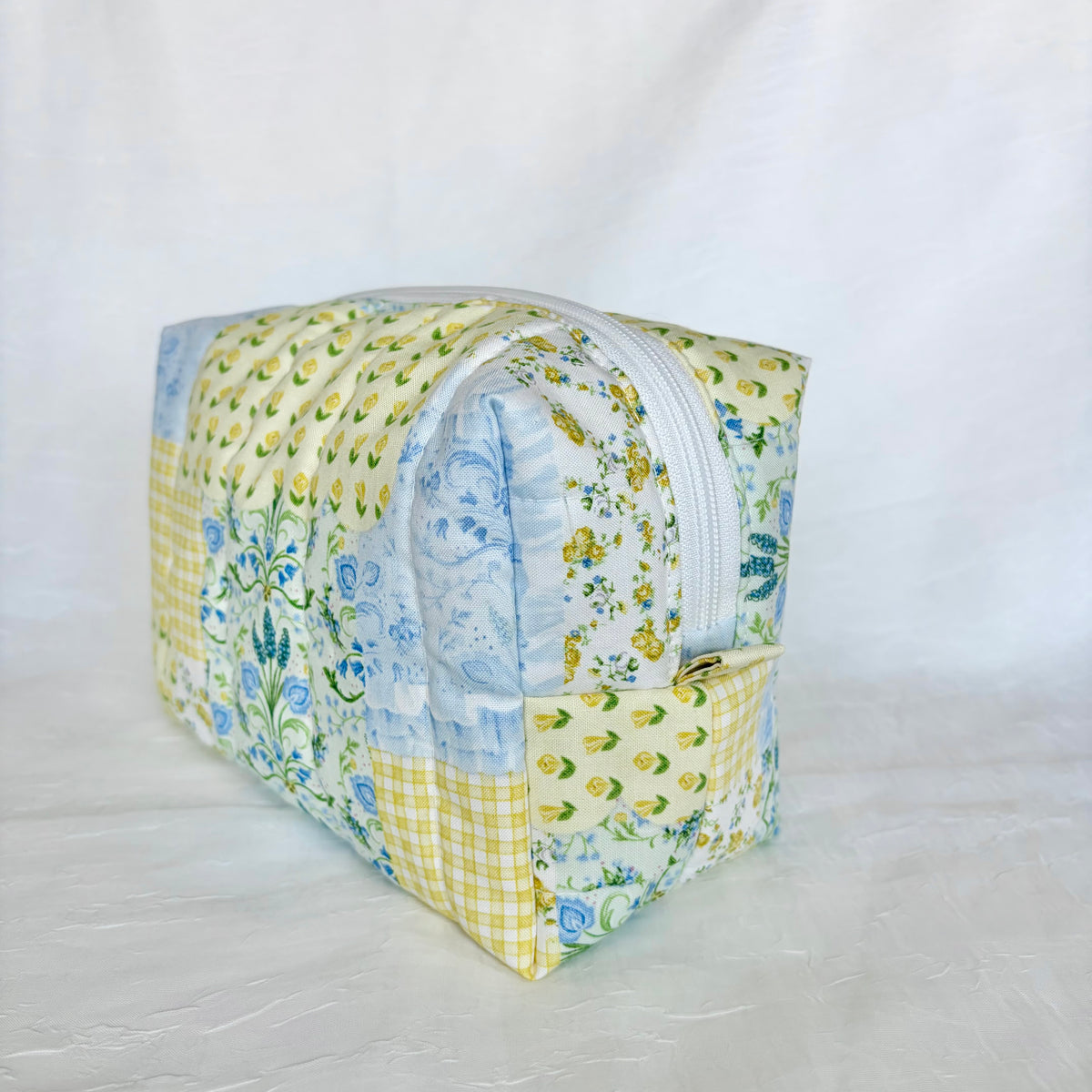 A handmade patchwork quilted zippered pouch with a floral and gingham pattern in yellow, blue, and white, set against a white fabric backdrop.