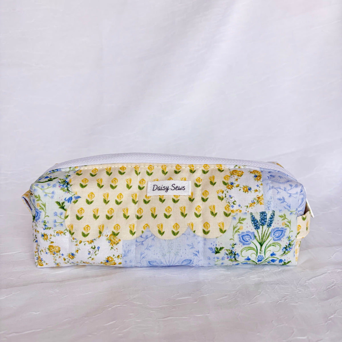 A fabric zippered pouch with a patchwork floral print in yellow, blue, and green on a white background, featuring a small white label that reads 'Daisy Sews'.
