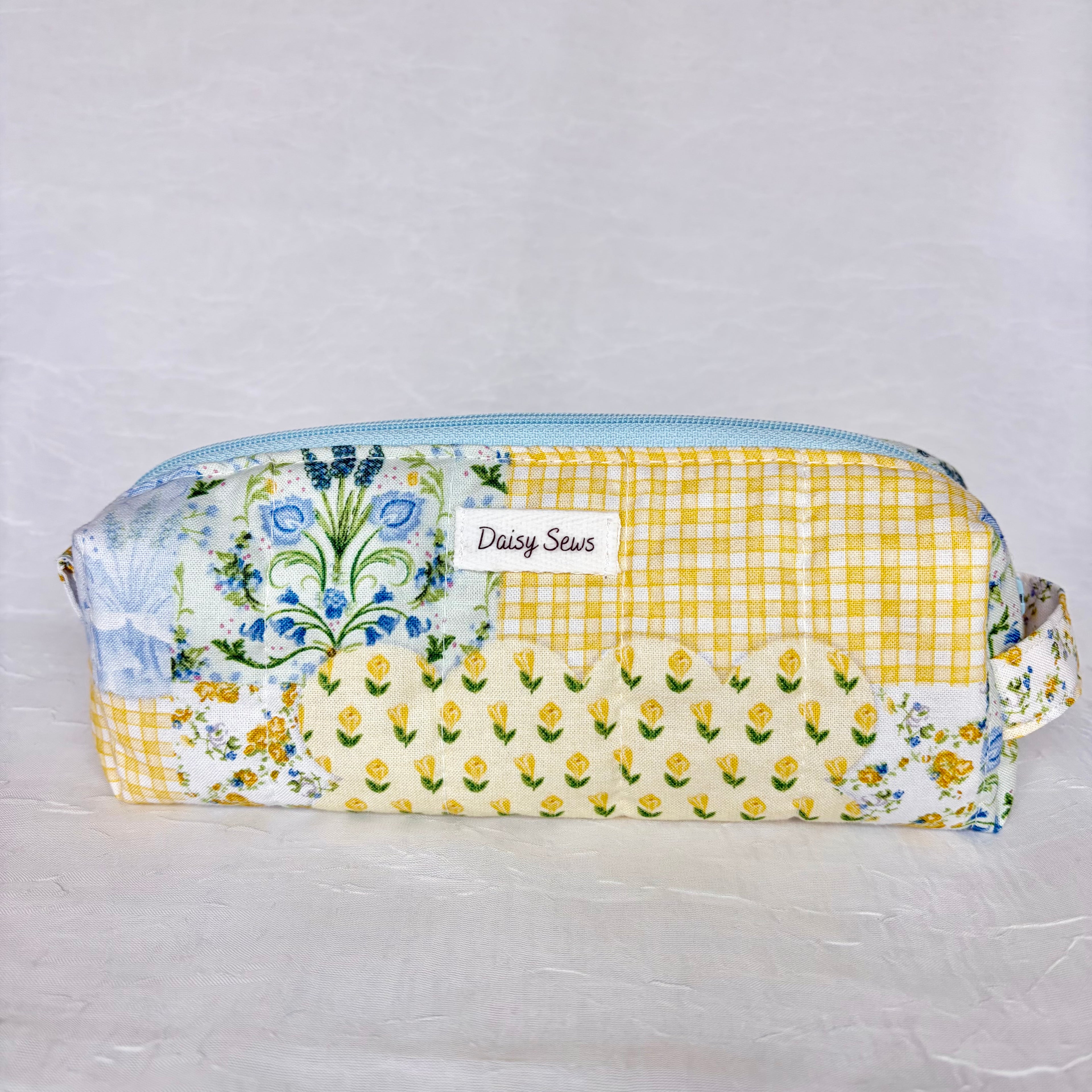 Blue and yellow floral patchwork boxy pencil bag with light blue zipper and a white label on white background