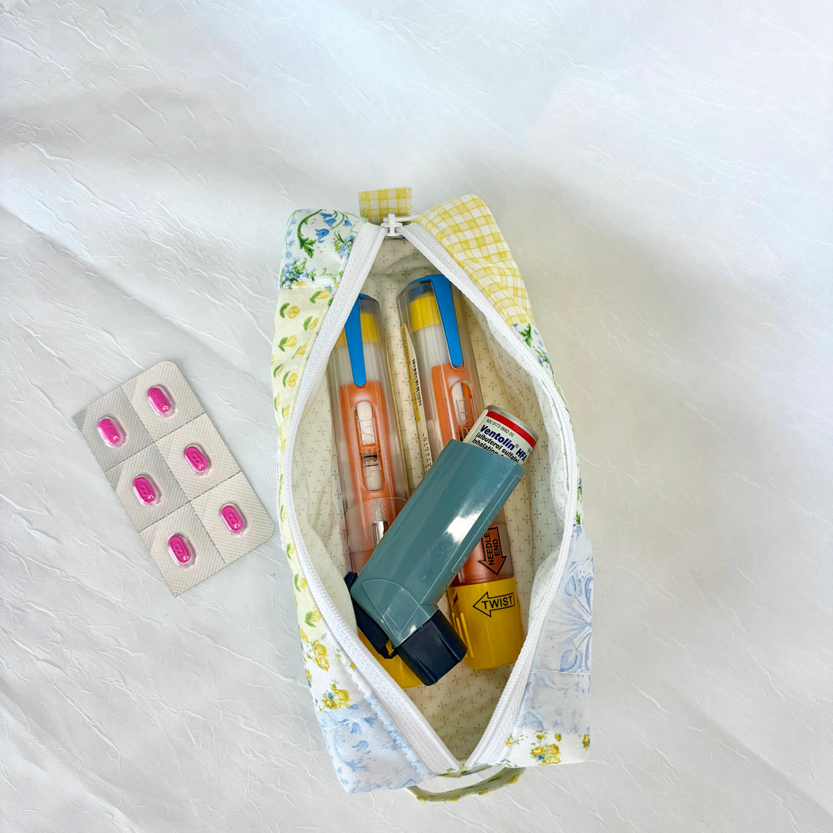 A floral zippered pouch filled with personal items, including an epipen, inhaler, and a blister pack of pink pills, laid on a white textured fabric background.